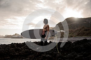 Side view brunette man standing in the black swimsuit with naked torso with a white surf in his hands on the shore