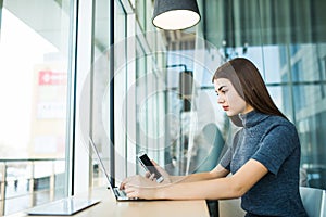 Side view of brunette woman using laptop computer sitting against big window in office