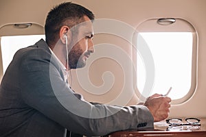 side view of businessman in earphones using smartphone in private jet.