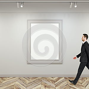Side view of businessman walking in modern gallery interior with empty white mock up poster frame on concrete wall and wooden