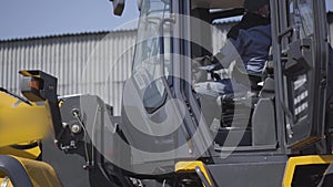 Side view of Caucasian driver sitting in cabin of yellow loader. Young employee in uniform inside heavy industrial