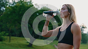 Side view of the caucasian female runner standing in the park, drinking water from the sport bottle, feeling thirsty