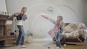 Side view of Caucasian girl in casual clothes fighting with pillows with younger siblings. Happy girls and boy playing