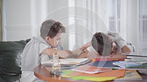 Side view of Caucasian schoolboy sitting at the table and writing down. His exhausted twin brother lying head on