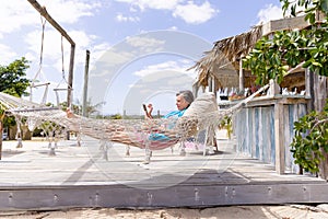 Side view of caucasian senior woman using digital tablet and resting on hammock at beach against sky