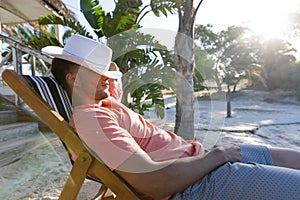 Side view of caucasian young man with hat on face sitting on deck chair against trees at beach