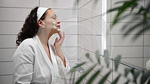 Side view of a charming African American pretty woman applying cleansing exfoliating beauty product on her face, looking at her re