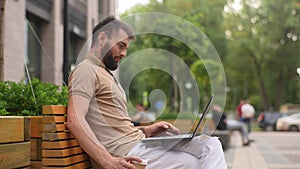 Side view of cheerful bearded freelancer male working on break with laptop and cup of coffee outdoors. Confide young