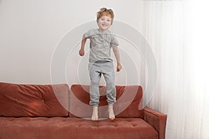Side view of cheerful boy jumps on couch while having fun at home. Home holidays.Blon hair happy boy in grey t-skirt and