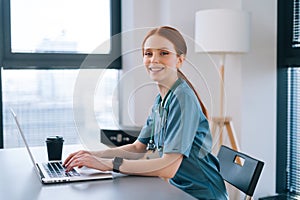 Side view of cheerful young female doctor in blue green medical uniform working typing on laptop keyboard sitting at