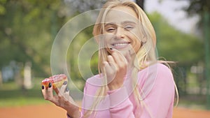 Side view of cheerful young woman tasting sweet donut and laughing. Portrait of charming blond Caucasian lady eating