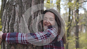 Side view close-up of cheerful brunette Caucasian girl approaching to tree in forest and hugging trunk. Pretty smiling
