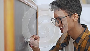 Side view close-up of happy smiling Middle Eastern guy in eyeglasses writing with chalk on blackboard indoors. Male
