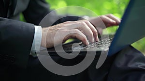 Side view close-up of male caucasian hands typing on laptop keyboard in summer park. Unrecognizable confident