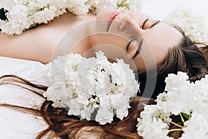 Side view close up portrait of cute millennial girl with fresh spring flowers, lying on cozy bed. Face of young beautiful brunette