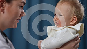 Side view close-up of smiling Caucasian female doctor talking in slow motion to unsatisfied infant. Baby girl crying as