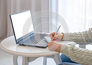 Side view and close up of woman`s hands using laptop with blank white screen and writing in notepad. Mockup laptop. Hands at work