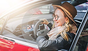 Side view, close up. Young smiling woman in glasses and hat sits behind wheel in red car. Trip, caravanning, tourism.