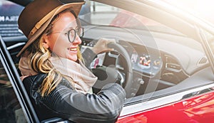 Side view, close up. Young smiling woman in glasses and hat sits behind wheel in red car. Trip, caravanning, tourism.