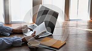 Side view of college student is reading book to find information and using laptop on wooden table.