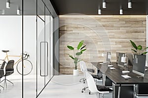 Side view on comfortable work places in stylish open space office with wooden wall, black and white furniture, glass doors and