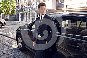 Side view of confident business man in formalwear and eyewear leaning at the car and looking at away