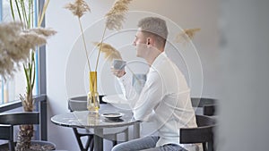 Side view of confident Caucasian businessman sitting at the table in cafe and drinking coffee. Relaxed young man in