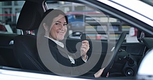 Side view of confident Caucasian woman sitting on driver`s seat in new car, showing keys at camera and smiling