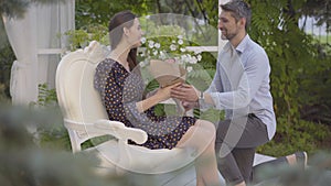 Side view of confident gorgeous woman sitting on white chair in pavilion as loving man approaching with bouquet of