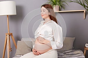 Side view of confident healthy caucasian pregnant woman with bare belly wearing white clothing sitting in bed in light bedroom