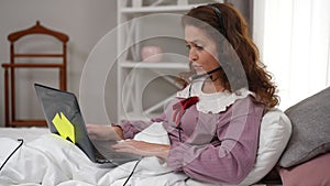 Side view confident smart mature woman in headphones talking surfing Internet lying in bed indoors. Intelligent