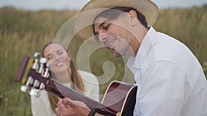 Side view confident talented young man playing guitar singing with blurred woman sitting at background on picnic. Happy