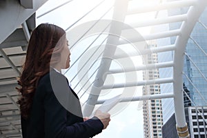 Side view of confident young Asian business woman with note standing and looking to forward far away at urban building city with c