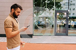 Side view of confident young man in casual clothes walking on city street, using mobile phone and drinking takeaway