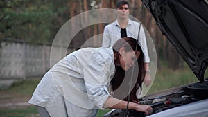 Side view confident young woman repairing automobile leaning at open car hood with blurred man talking at background