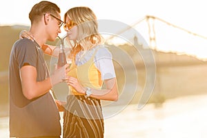 side view of couple drinking beverage with two plastic straws from one bottle on river beach