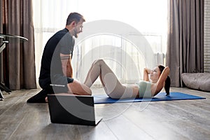 side view on couple working out at home, man helping woman doing sit-ups crunches on mat in living room