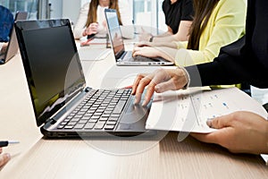 Side view of crop female working on laptop and taking notes on paper while sitting at table with group of colleagues in