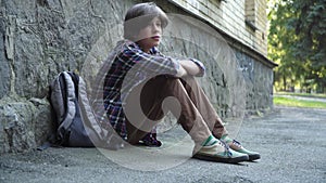 Side view of crying bullied schoolboy sitting outdoors at rock wall. Wide shot of sad Caucasian lonely boy with backpack