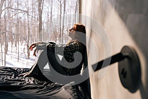 Side view of crying young woman sitting by window on sunny day on windowsill with winter nature background, feels upset