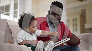 Side view curios African American son enjoying leisure with man reading book. Cute hyperactive toddler boy listening
