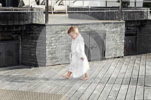 Side view of cute funny caucasian blond kid boy wearing white pool spa bathrobe scolded walking by tiled stone floor