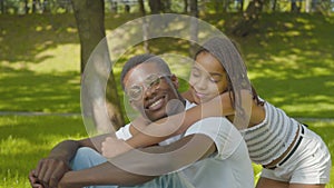 Side view of cute girl hugging man in sunny summer park. Portrait of happy African American sister and brother looking