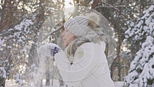 Side view cute girl in warm winter clothes throws snow up and laughs cheerfully. Beautiful blurred background of snowy