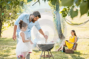 Girl watching father preparing meat on barbecue grill during family picnic