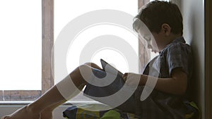 SIDE VIEW: Cute little boy uses a white tablet PC on a windowsill at home