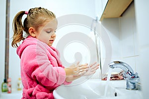 Side view of cute little girl with ponytail in pink bathrobe washing her hands.