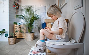 Side view of cute small boy sitting on toilet indoors at home, using smartphone.