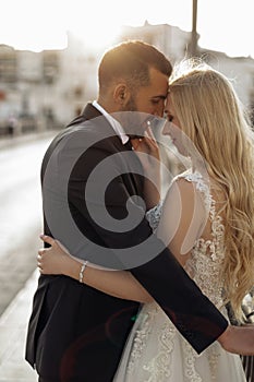 Side view of cute wedding couple standing on street with closed eyes. Young woman bride embracing with groom in suit.