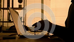 Side view dark silhouette of a man typing on a laptop keyboard. A male manager, designer or programmer in a black shirt